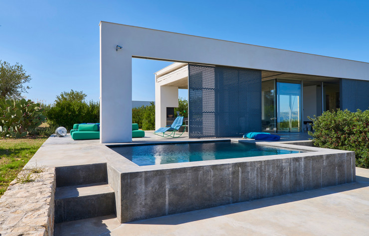 Habillée de carrelage en pierre de lave, la piscine s’ouvre sur le paysage vers la mer. Un ensemble de pare-soleil mobiles court le long des baies vitrées et jusque sur la terrasse, servant aussi de coupe-vent.