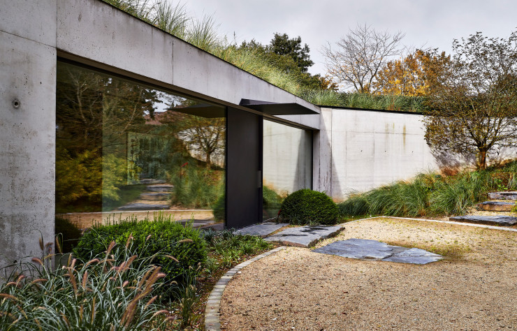 Au seuil de la maison, le béton des parements a été laissé brut volontairement, comme pour l’ensemble du bâtiment. L’effet miroir des grandes baies vitrées accentue la présence de la végétation.
