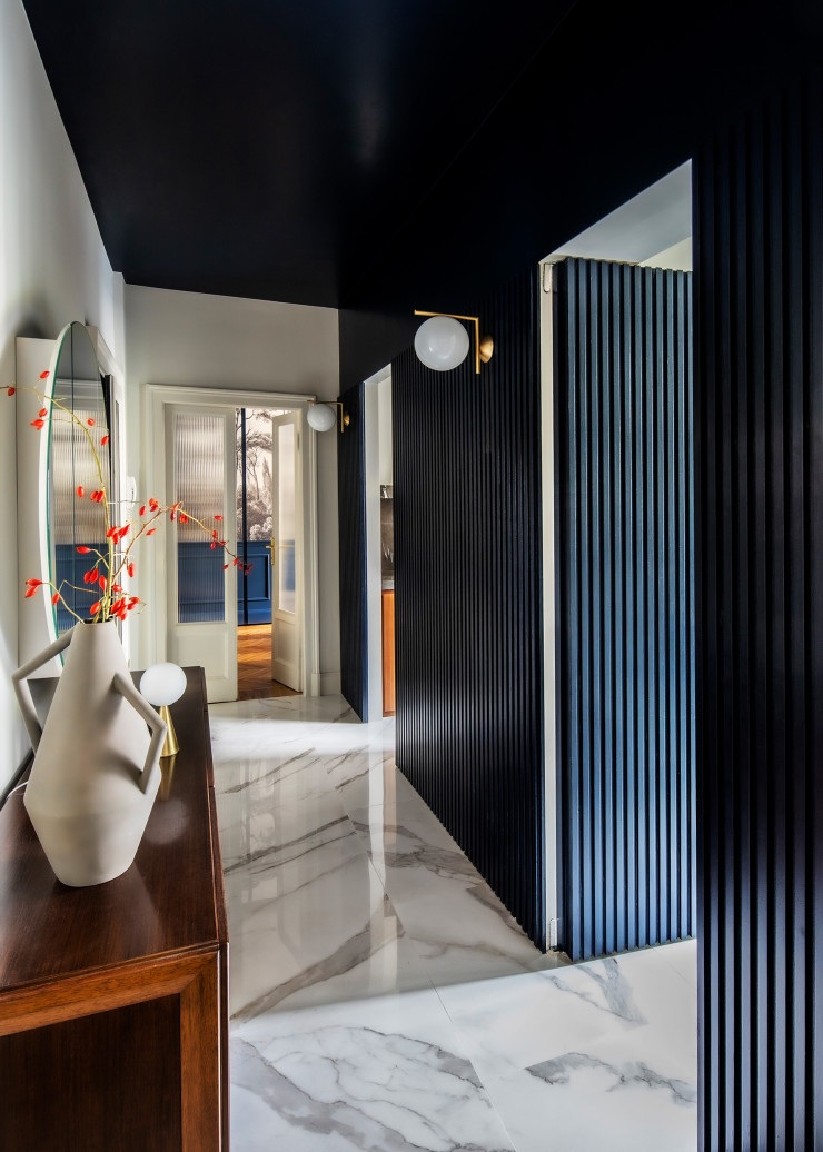 Le couloir se pare d’un lambris en bois teinté au fraisage vertical, conçu par Andrea Rubini, ce dernier étant également à l’origine du miroir rond en triptyque ajustable. Applique IC Lights, design Michael Anastassiades (Flos). Sur le buffet vintage, de Franco Albini (Poggi), vase Kora, design Studiopepe (Atipico), et lampe Bilia Mini, design Gio Ponti (Fontana Arte). Sol en grès cérame Dreaming (Lea Ceramiche).