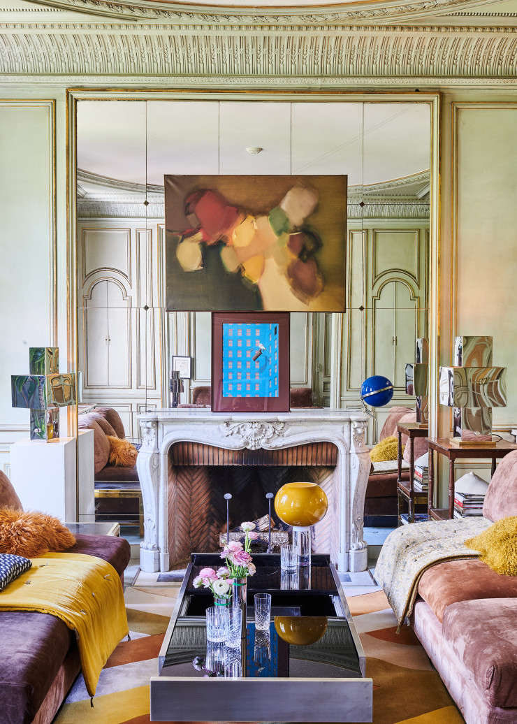 La convivialité était de rigueur chez Willy Rizzo. Dans le grand salon, la table basse en verre et en acier s’ouvre en deux, révélant un bar dissimulé dans le pied. Les deux canapés C sont modulables sans limites de taille et n’ont pas d’accoudoirs pour accueillir un maximum de convives. Dans le reflet du miroir, on aperçoit à droite la bulle bleue d’une Galileo, la console HI-FI conçue par le designer pour écouter de la musique en y branchant un iPad ou un iPhone. Au-dessus de la cheminée, Le Suicide de Freud, de Jean Lagarrigue, sous une photo de Suzy Parker, mannequin puis actrice, dans l’escalier de Mademoiselle Chanel, en 1956. Sur la table basse, lampe Bud 3 en verre soufflé et en PMMA (un polymère), design Rodolfo Dordoni (Foscarini). Tapis Laminae Anigre en laine et en soie mate, collection « Bloom », design Jeff Leatham (Tai Ping). Jetés de canapés Velours lavé (à gauche) en velours lavé et Imprimé (à droite) en velours lavé et en coton (Caravane). Dessus, plaid Mascara en laine et en polyamide, design Moisés Hernández (Ligne Roset). Coussins en lin et en peau de chèvre du Tibet (Maison de Vacances).