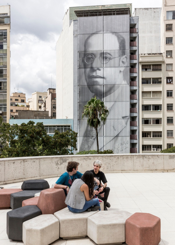Vue sur l’architecture brutaliste du Centro, décorée du portrait XXL de l’artiste Mário de Andrade (reproduisant une photo de la série « Giganto », de Raquel Brust) depuis la terrasse de la bibliothèque Mário de Andrade. Au premier plan, sièges Campo en béton teinté, un des best-sellers de la marque brésilienne Ovo, que l’on retrouve également à la pinacothèque.